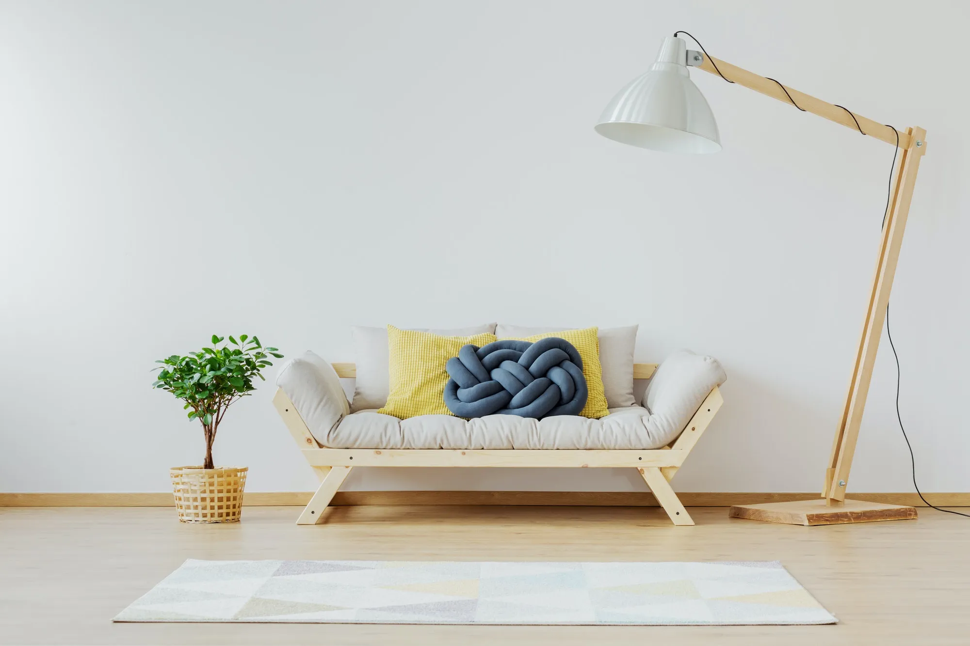 A minimalist living room featuring a wooden-framed sofa with cozy cushions, a large white lamp, and a small potted plant in the corner.