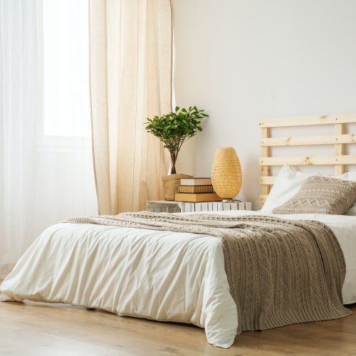 Simple Cozy Bedroom with Natural Elements A cozy bedroom with a wooden bed frame, white linens, and a beige throw blanket. The room features natural light, a potted plant, a stack of books, and a decorative woven lamp.